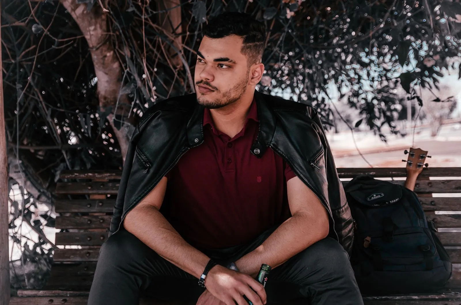 A thoughtful man in a leather jacket sits on a bench outdoors with a backpack and guitar.